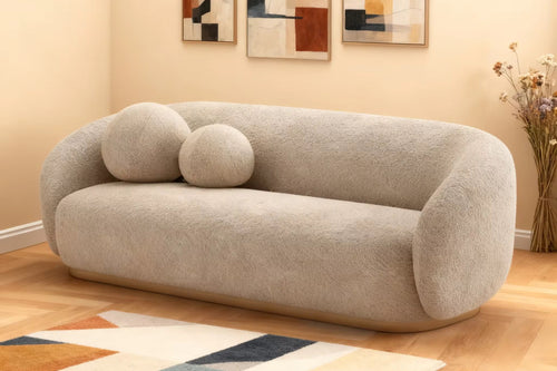 A modern curved sofa in a textured light beige boucle fabric, styled in a warm living room setting with two matching round throw pillows, a geometric patterned rug, and abstract wall art.