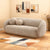 A modern curved sofa in a textured light beige boucle fabric, styled in a warm living room setting with two matching round throw pillows, a geometric patterned rug, and abstract wall art.