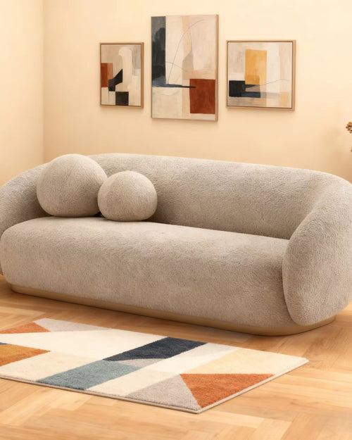 A modern curved sofa in a textured light beige boucle fabric, styled in a warm living room setting with two matching round throw pillows, a geometric patterned rug, and abstract wall art.
