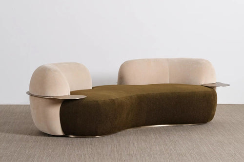 A modern curved lounge chair with a dark olive green velvet seat and light beige velvet arms and backrest, featuring thin metallic side tables attached to the arms, set against a minimalist studio background.