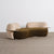 A modern curved lounge chair with a dark olive green velvet seat and light beige velvet arms and backrest, featuring thin metallic side tables attached to the arms, set against a minimalist studio background.