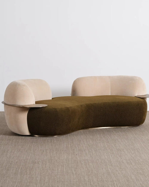 A modern curved lounge chair with a dark olive green velvet seat and light beige velvet arms and backrest, featuring thin metallic side tables attached to the arms, set against a minimalist studio background.