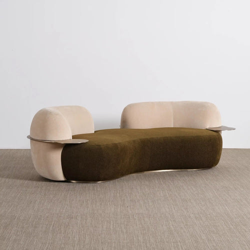 A modern curved lounge chair with a dark olive green velvet seat and light beige velvet arms and backrest, featuring thin metallic side tables attached to the arms, set against a minimalist studio background.