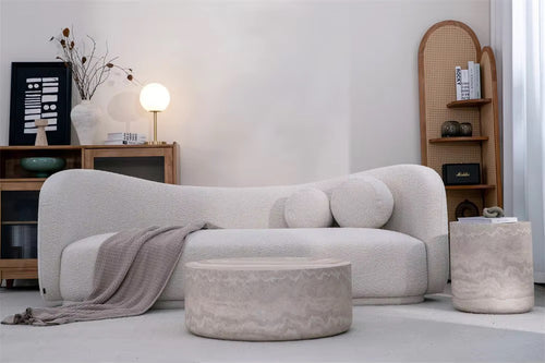 A modern curved upholstered sofa in a light warm gray or off-white bouclé fabric, styled in a minimalist living room with a matching ottoman, a side table, and wooden shelving.