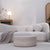 A modern curved upholstered sofa in a light warm gray or off-white bouclé fabric, styled in a minimalist living room with a matching ottoman, a side table, and wooden shelving.