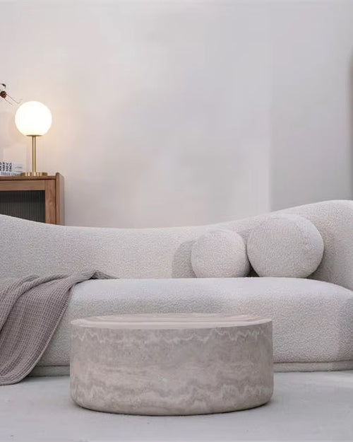 A modern curved upholstered sofa in a light warm gray or off-white bouclé fabric, styled in a minimalist living room with a matching ottoman, a side table, and wooden shelving.