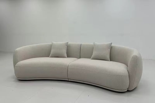 A curved upholstered sofa in a light warm gray or off-white fabric, shown in a minimalist showroom with white walls and ceiling tiles.