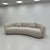 A curved upholstered sofa in a light warm gray or off-white fabric, shown in a minimalist showroom with white walls and ceiling tiles.