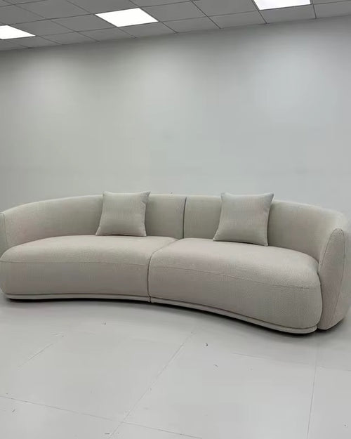 A curved upholstered sofa in a light warm gray or off-white fabric, shown in a minimalist showroom with white walls and ceiling tiles.