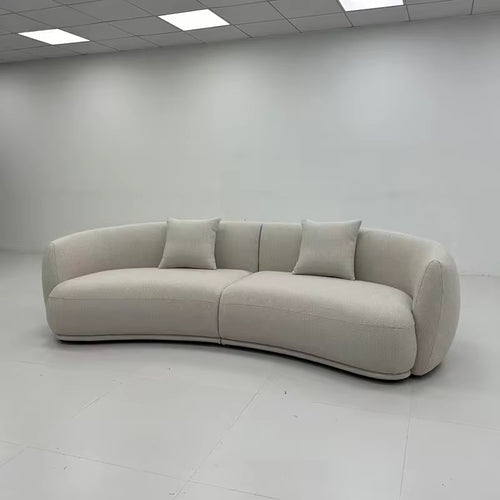 A curved upholstered sofa in a light warm gray or off-white fabric, shown in a minimalist showroom with white walls and ceiling tiles.