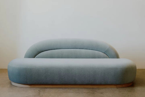 Gemini Couch featuring blue upholstered seating, a low-profile organic silhouette, rounded arms, a gently curved backrest, and a subtle wooden base.