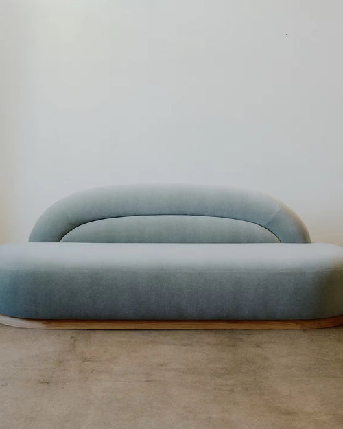 Gemini Couch featuring blue upholstered seating, a low-profile organic silhouette, rounded arms, a gently curved backrest, and a subtle wooden base.