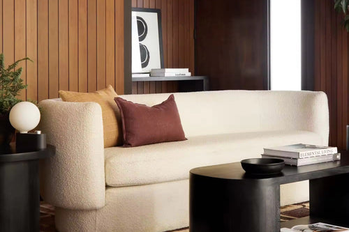 A cream bouclé sofa in a modern living room setting with wood-paneled walls, a patterned rug, a black coffee table, and accent pillows.
