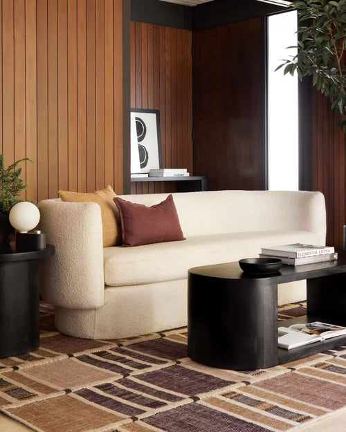 A cream bouclé sofa in a modern living room setting with wood-paneled walls, a patterned rug, a black coffee table, and accent pillows.