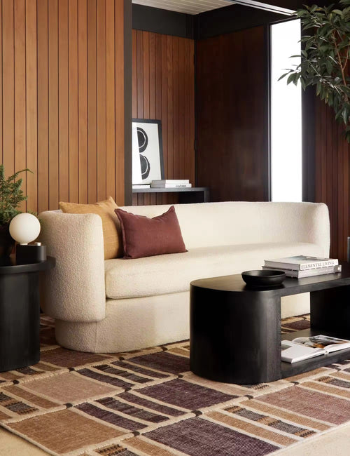 A cream bouclé sofa in a modern living room setting with wood-paneled walls, a patterned rug, a black coffee table, and accent pillows.