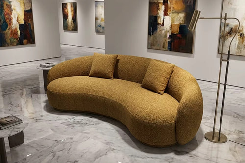 A curved, low-profile boucle sofa in a warm mustard-yellow or ochre color, styled in a modern art gallery with marble floors and abstract paintings on the walls.
