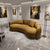 A curved, low-profile boucle sofa in a warm mustard-yellow or ochre color, styled in a modern art gallery with marble floors and abstract paintings on the walls.