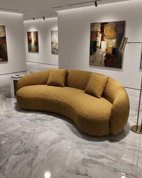 A curved, low-profile boucle sofa in a warm mustard-yellow or ochre color, styled in a modern art gallery with marble floors and abstract paintings on the walls.