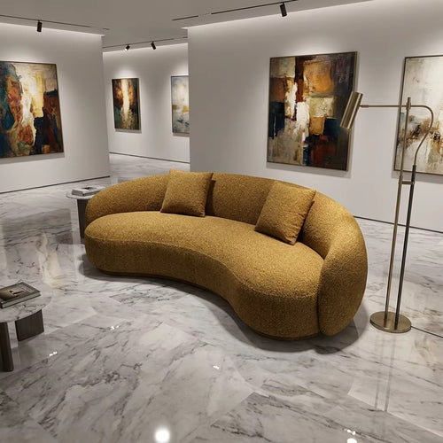 A curved, low-profile boucle sofa in a warm mustard-yellow or ochre color, styled in a modern art gallery with marble floors and abstract paintings on the walls.