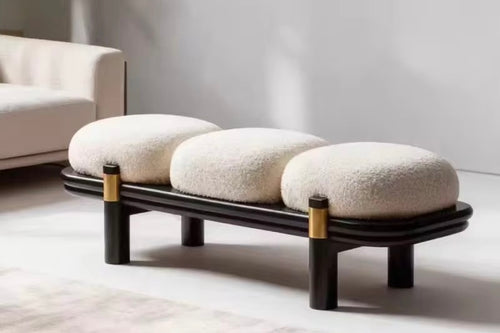 A modern ottoman online with three oval cream boucle cushions on a curved black wooden frame, accented with gold metal rings on the legs, set in a minimalist living room.