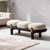A modern ottoman online with three oval cream boucle cushions on a curved black wooden frame, accented with gold metal rings on the legs, set in a minimalist living room.