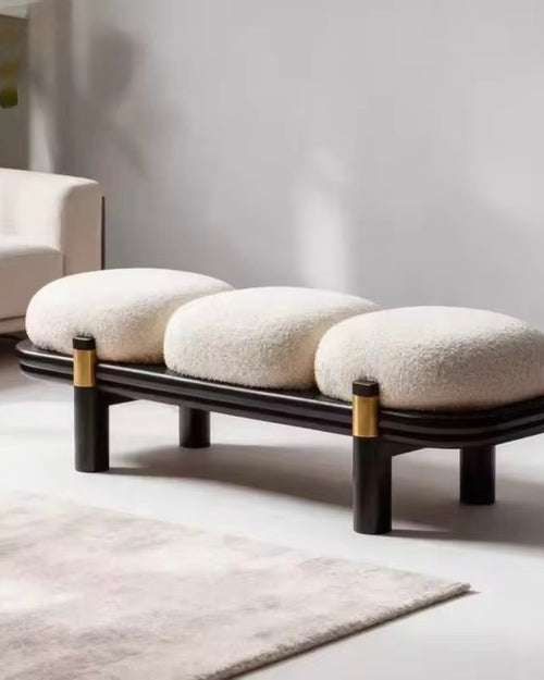 A modern ottoman online with three oval cream boucle cushions on a curved black wooden frame, accented with gold metal rings on the legs, set in a minimalist living room.