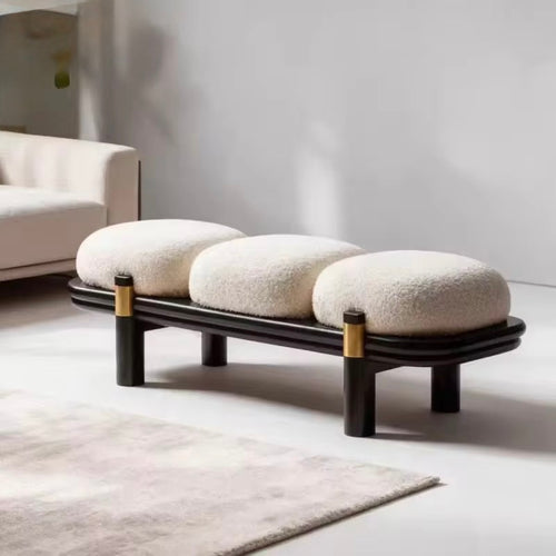 A modern ottoman online with three oval cream boucle cushions on a curved black wooden frame, accented with gold metal rings on the legs, set in a minimalist living room.