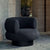 A modern round armchair upholstered in dark charcoal bouclé fabric with a black cylindrical base, set against a stone architectural background.
