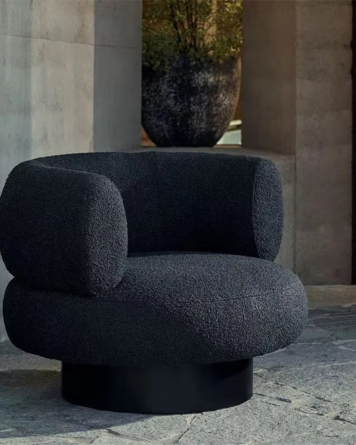 A modern round armchair upholstered in dark charcoal bouclé fabric with a black cylindrical base, set against a stone architectural background.