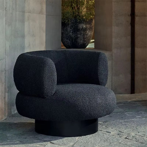 A modern round armchair upholstered in dark charcoal bouclé fabric with a black cylindrical base, set against a stone architectural background.